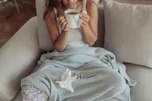 простуженная девушка сидит с чашкой чая и под пледом, на нем носовые платочки и таблетки